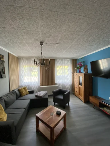 Wohnzimmer mit Sofa, Sessel, Holztisch, Fernseher und hellen Fenstern.