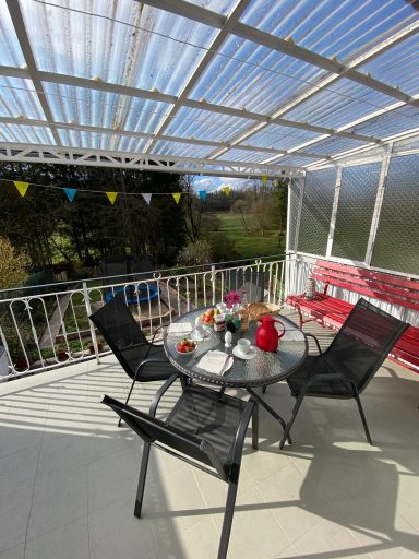 Tisch mit Stühlen auf einer überdachten Terrasse, umgeben von Pflanzen und Blick ins Grüne.