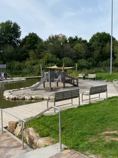 Spielplatz am Wasser mit Bänken und Spielgeräten, umgeben von Bäumen.