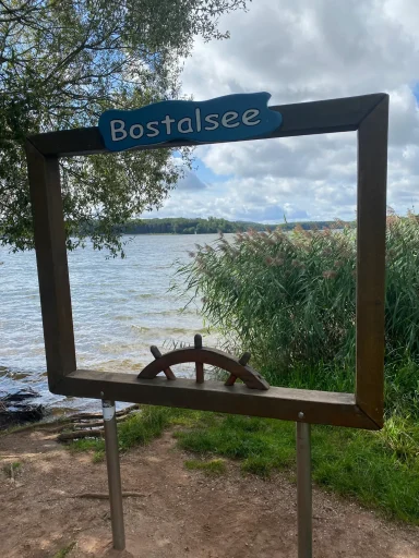 Holzrahmen mit Schriftzug „Bostalsee“ und Steuerrad am Ufer eines Sees.