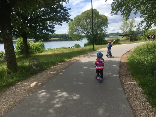 Zwei Kinder fahren mit Scootern auf einem Weg am See, umgeben von Bäumen und Gras.